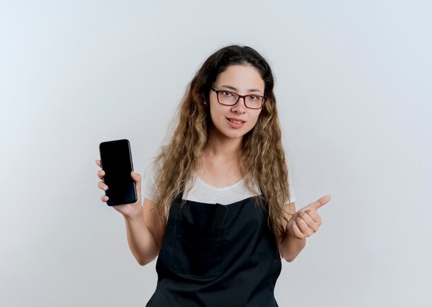 Junge professionelle Friseurfrau in der Schürze, die das lächelnde Smartphone zeigt, das vorne zeigt, zeigt Daumen hoch, die über weiße Wand stehen