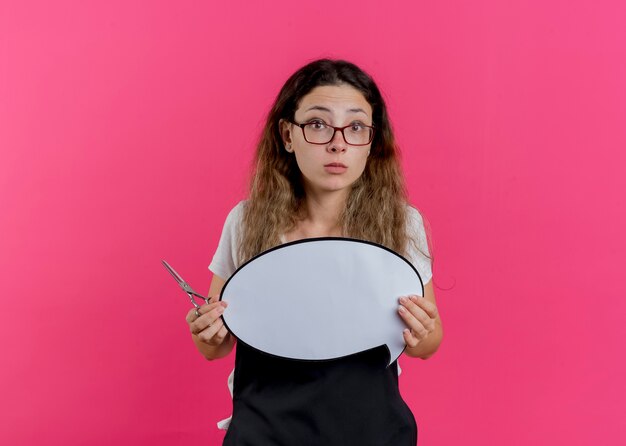 Junge professionelle friseurfrau in der schürze, die schere und leeres sprachblasenzeichen hält, das vorne betrachtet wird, das über rosa wand steht