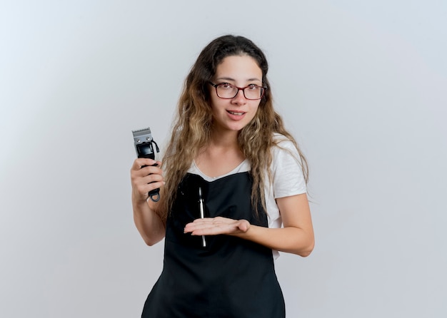 Junge professionelle Friseurfrau in der Schürze, die Trimmer hält, der es mit Arm ihrer Hand lächelnd steht über weißer Wand präsentiert