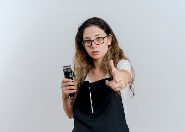 Junge professionelle Friseurfrau in der Schürze, die Trimmer hält, der mit Zeigefinger an der Vorderseite zeigt und zuversichtlich steht über weißer Wand