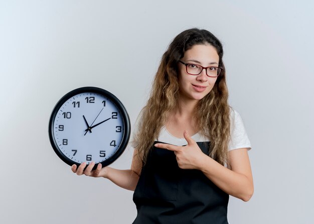 Junge professionelle Friseurfrau in der Schürze, die Wanduhr hält, die mit dem Zeigefinger darauf lächelt und über weißer Wand steht