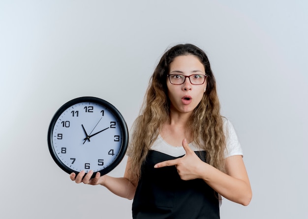 Junge professionelle Friseurfrau in der Schürze, die Wanduhr Pointign mit Zeigefinger hält, der überrascht ist, über weißer Wand zu stehen