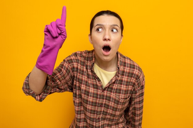 Junge Putzfrau im karierten Hemd in Gummihandschuhen, die fasziniert und überrascht aussieht und Zeigefinger mit weit geöffnetem Mund über oranger Wand steht