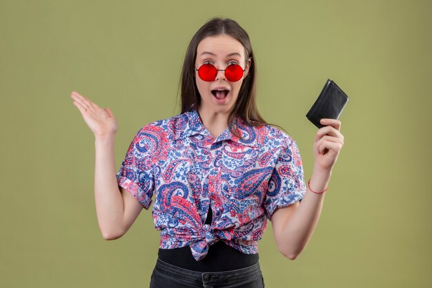 Junge reisende Frau, die rote Sonnenbrille hält, die Brieftasche hält, die erstaunt und überrascht mit erhobenen Armen steht, die über grünem Hintergrund stehen