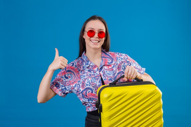 Junge reisende Frau, die rote Sonnenbrille hält, die gelben Koffer hält, der fröhlich Daumen über blaue Wand zeigt