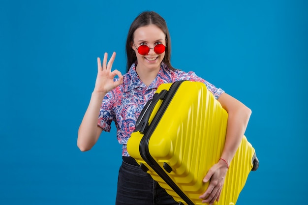 Junge reisende Frau, die rote Sonnenbrille hält, die gelben Koffer hält, der Kamera positiv und glücklich lächelnd tut, das ok Zeichen steht über blauem Hintergrund