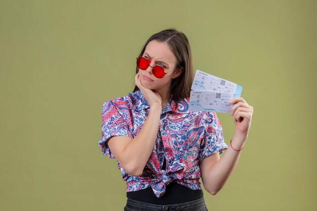 Junge Reisende Frau, die rote Sonnenbrille hält, die Tickets hält, die mit Hand auf Kinn unzufrieden stehen und mit nachdenklichem Ausdruck über grünem Hintergrund denken