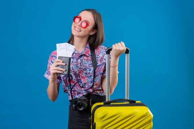 Junge reisende Frau, die rote Sonnenbrille mit gelbem Koffer hält, der Pass und Tickets hält, die fröhlich mit glücklichem Gesicht über blauer Wand lächeln