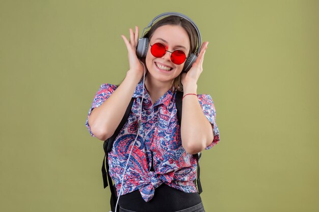 Junge reisende frau, die rote sonnenbrille trägt und mit rucksack musik hört, die kopfhörer verwendet, die mit glücklichem gesicht über grüner wand lächeln