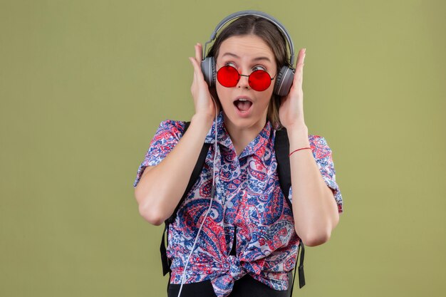 Junge reisende Frau, die rote Sonnenbrille trägt und mit Rucksack Musik hört, die Kopfhörer verwendet, die über grüner Wand überrascht schauen
