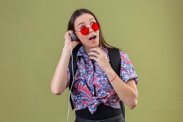 Junge reisende Frau, die rote Sonnenbrille trägt und mit Rucksack Musik hört, die Kopfhörer verwendet, die überrascht und glücklich stehen über grünem Hintergrund stehen