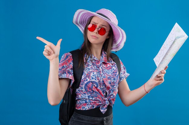 Junge reisende Frau im Sommerhut, der rote Sonnenbrille hält Karte hält, die mit nachdenklichem Ausdruck beiseite schaut, der mit dem Finger zur Seite denkt und Zweifel hat, die über blauem Hintergrund stehen