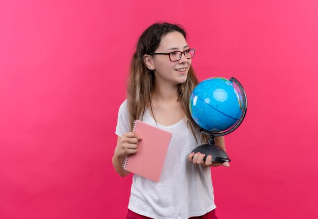 Junge reisende Frau im weißen T-Shirt, das Globus und Notizbuch hält, das positiv und glücklich lächelnd über rosa Wand beiseite schaut