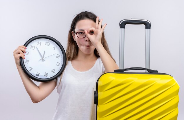 Junge reisende Frau im weißen T-Shirt, das mit Koffer hält Wanduhr tut ok Zeichen, das durch dieses Zeichen über weiße Wand schaut