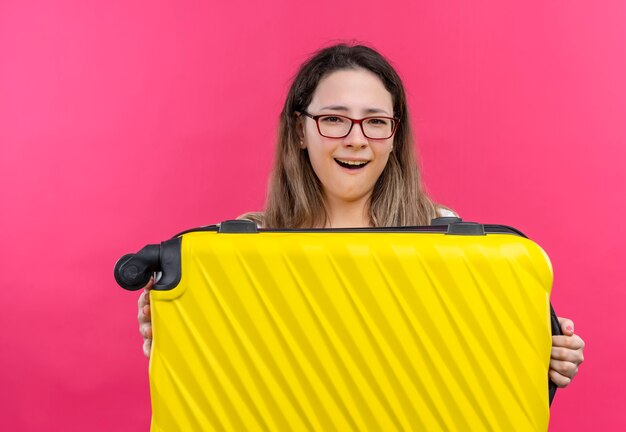 Junge reisende Frau im weißen T-Shirt, das Reisekoffer hält, der fröhlich über rosa Wand steht