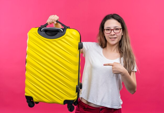 Junge reisende Frau im weißen T-Shirt, das Reisekoffer hält, der mit Zeigefinger auf ihn lächelt, der zuversichtlich über rosa Wand steht