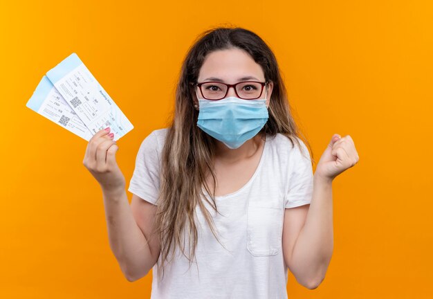 Junge reisende Frau im weißen T-Shirt, das schützende Gesichtsmaske hält, die Flugkarten zusammenhält, die aufgeregte Faust und geballtes Stehen über orange Wand ballen