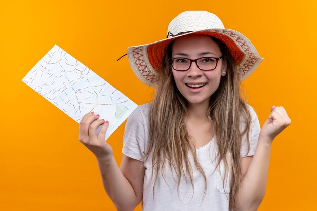 Junge reisende Frau im weißen T-Shirt, das Sommerhut hält, der Karte zusammenballt Faust aufgeregt und glücklich lächelnd steht über orange Wand