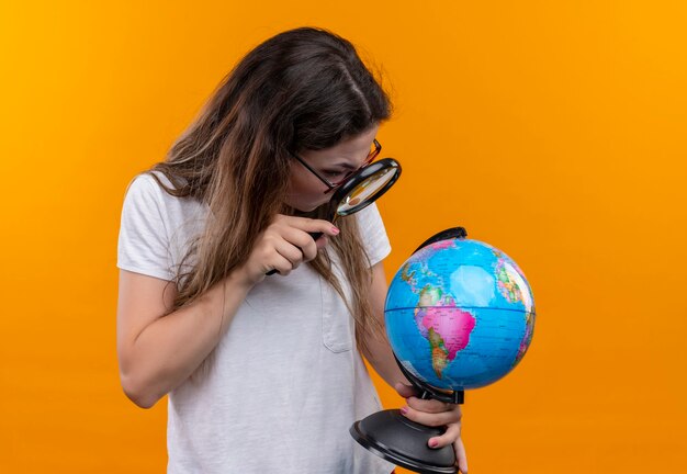 Kostenloses Foto junge reisende frau im weißen t-shirt hält globus, der sie durch lupe betrachtet, die überrascht steht, über orange wand stehend