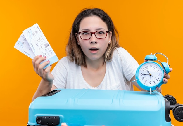 Kostenloses Foto junge reisende frau im weißen t-shirt stehend mit koffer voll von kleidern, die flugtickets und wecker halten, die über orange wand überrascht und erstaunt schauen