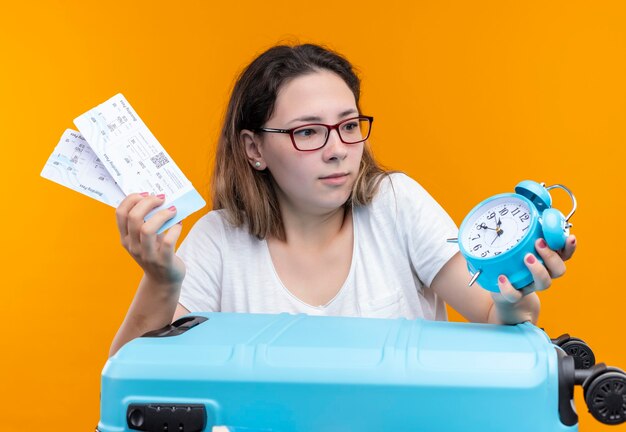 Junge Reisende Frau im weißen T-Shirt stehend mit Koffer voll von Kleidern, die Flugtickets und Wecker halten, die über orange Wand überrascht und erstaunt schauen