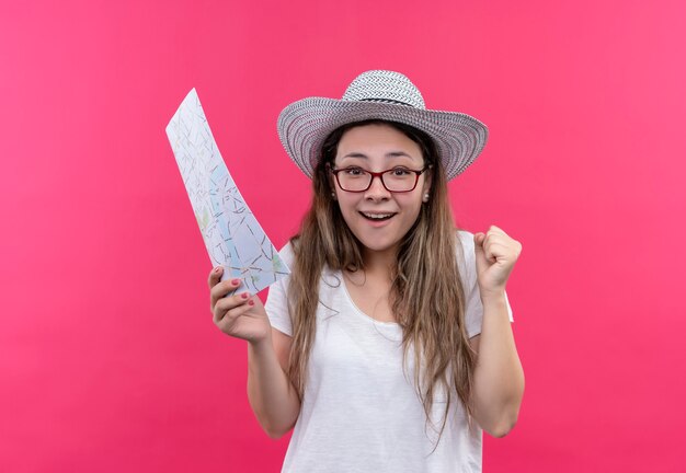 Junge reisende Frau im weißen T-Shirt und im Sommerhut, die Karte halten, die aufgeregt und glückliche geballte Faust steht, die über rosa Wand steht