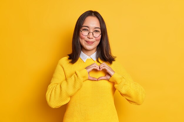 Junge romantische asiatische Frau mit zartem Gesichtsausdruck formt Herzgeste drückt Liebe zum Freund aus, trägt runde Brille und Strickpullover.