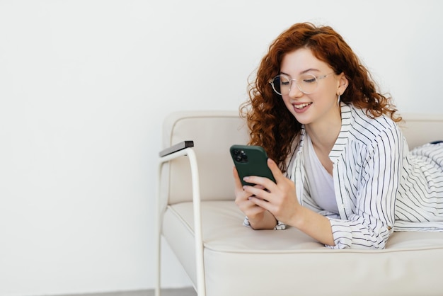 Junge rothaarige Frau mit Smartphone, während sie sich zu Hause auf dem Sofa ausruht