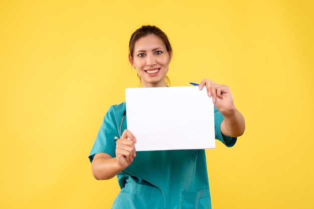 Junge Ärztin der Vorderansicht im medizinischen Hemd, die Papieranalyse auf gelbem Hintergrund hält