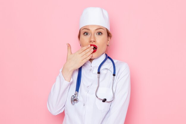 Junge Ärztin der Vorderansicht im weißen Anzug mit blauem Stethoskop, das auf der Emotion der rosa Raumjobfrau aufwirft