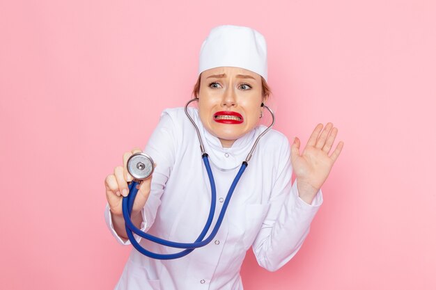 Junge Ärztin der Vorderansicht im weißen Anzug mit blauem Stethoskop, das auf der rosa Raumweibchenarbeit aufwirft und misst