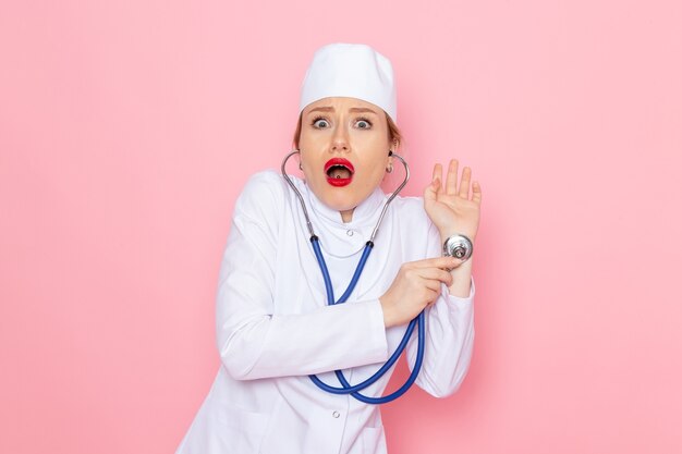 Junge Ärztin der Vorderansicht im weißen Anzug mit blauem Stethoskop, das die Messung mit überraschtem Ausdruck auf dem medizinischen Krankenhaus der rosa Raummedizin darstellt