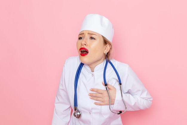 Junge Ärztin der Vorderansicht im weißen Anzug mit blauem Stethoskop, das Herzschmerz auf der rosa Raumweibchenarbeit aufwirft