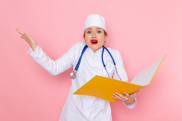 Junge Ärztin der Vorderansicht im weißen medizinischen Anzug mit blauem Stethoskop, das gelbe Dateien auf dem rosa Raum hält