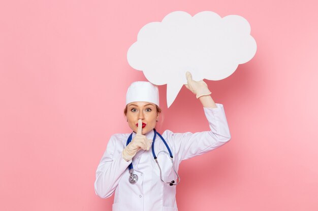 Junge Ärztin der Vorderansicht im weißen medizinischen Anzug mit blauem Stethoskop, das großes weißes Zeichen auf dem medizinischen Raumkrankenhaus der rosa Raummedizin hält