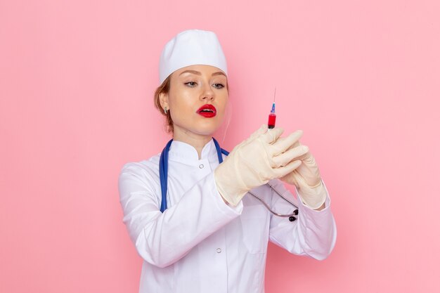 Junge Ärztin der Vorderansicht im weißen medizinischen Anzug mit blauem Stethoskop, das Injektion auf der rosa Raummedizinmedizinkrankenhausarbeiterin weiblich hält