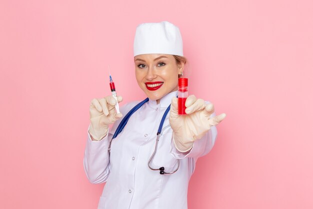 Junge Ärztin der Vorderansicht im weißen medizinischen Anzug mit blauem Stethoskop, das Injektion und Flasche auf der rosa Raummedizin-Krankenhausgesundheit hält