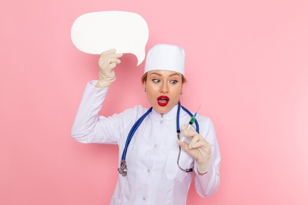 Junge Ärztin der Vorderansicht im weißen medizinischen Anzug mit blauem Stethoskop, das Injektion und weißes Zeichen auf der medizinischen Krankenhausgesundheit des rosa Raummedizin hält