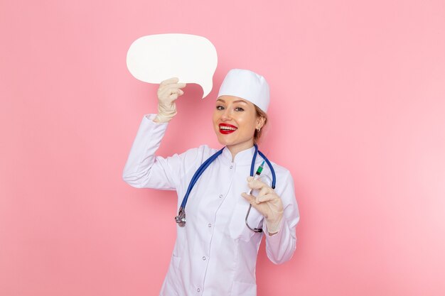 Junge Ärztin der Vorderansicht im weißen medizinischen Anzug mit blauem Stethoskop, das Injektion und weißes Zeichen hält, das auf dem rosa Raummedizin-medizinischen Krankenhausjob lächelt