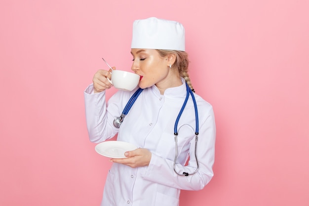 Junge Ärztin der Vorderansicht im weißen medizinischen Anzug mit blauem Stethoskop, das Kaffee auf dem medizinischen Krankenhaus der rosa Raummedizin trinkt