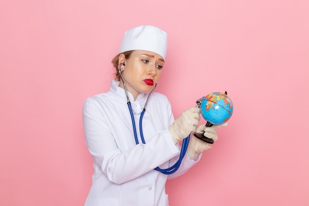 Junge Ärztin der Vorderansicht im weißen medizinischen Anzug mit blauem Stethoskop, das kleinen Globus auf der medizinischen Krankenhausarbeit der rosa Raummedizin prüft