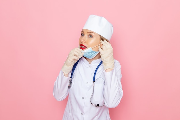 Junge Ärztin der Vorderansicht im weißen medizinischen Anzug mit blauem Stethoskop, das sterile Maske auf dem rosa Raum trägt