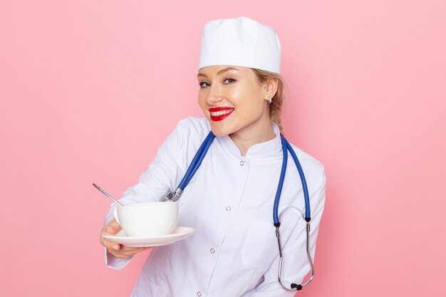 Junge Ärztin der Vorderansicht im weißen medizinischen Anzug mit blauem Stethoskop, das Tasse Kaffee lächelnd auf dem rosa Raummedizin-medizinischen Krankenhausjob hält