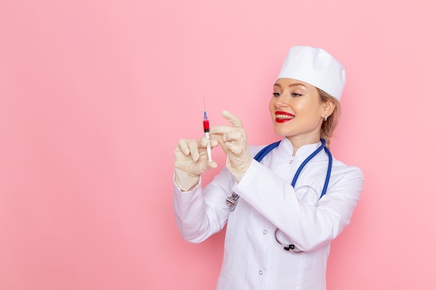 Junge Ärztin der Vorderansicht im weißen medizinischen Anzug mit dem blauen Stethoskop, das die Injektion hält, die auf der rosa Raummedizinmedizin-medizinischen Krankenhausarbeiterfrau lächelt