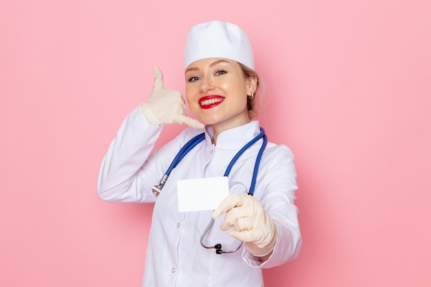 Junge Ärztin der Vorderansicht im weißen medizinischen Anzug mit dem blauen Stethoskop, das lächelnde weiße Karte hält, die auf dem rosa Raum aufwirft