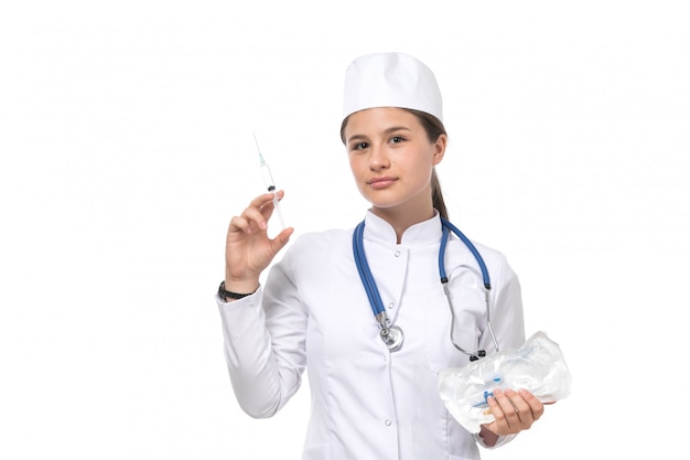 Junge Ärztin der Vorderansicht im weißen medizinischen Anzug und in der weißen Kappe mit blauem Stethoskop, das Injektion hält