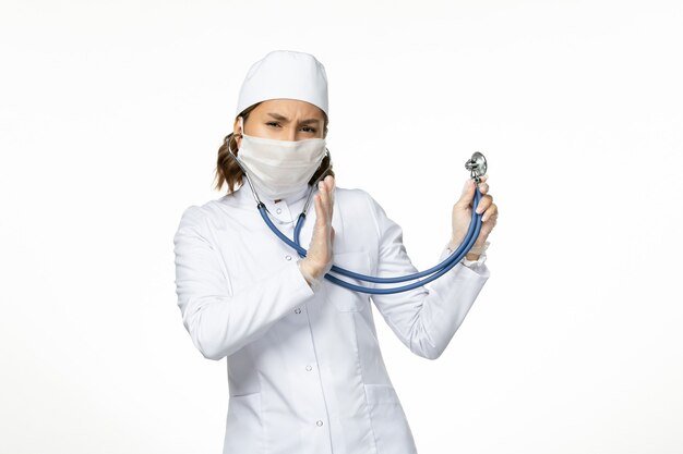 Junge Ärztin der Vorderansicht mit steriler Maske wegen Coronavirus, der Stethoskop auf weißem Schreibtisch hält