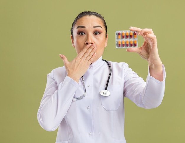 Junge Ärztin, die Blister mit Pillen hält, die schockiert sind und den Mund mit der Hand bedecken