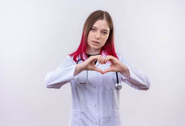 junge Ärztin, die das medizinische Gewand des Stethoskops trägt, das Herzgeste auf isolierter weißer Wand zeigt