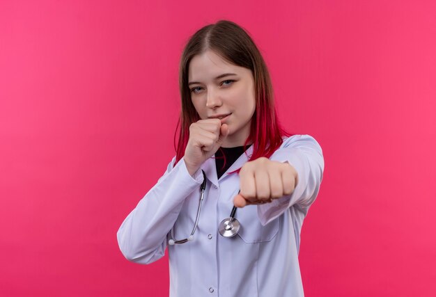 junge Ärztin, die das medizinische Kleid des Stethoskops trägt, das in der Kampfhaltung auf rosa isolierter Wand steht